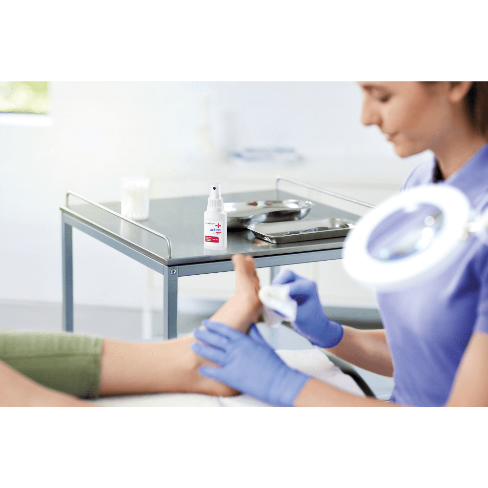 In una clinica, una persona con i guanti blu sta pulendo il piede di un’altra persona. L’attenzione è rivolta alla prevenzione delle infezioni. Sul tavolo sono presenti un flacone da 50 ml di Schülke octenisept® spray disinfettante per ferite della Schülke & Mayr GmbH e un vassoio di metallo con gli strumenti.