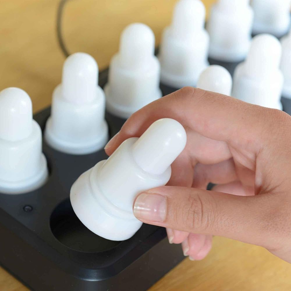 Una mano posiziona o rimuove una candela LED bianca e rotonda dal vassoio nero del set LED della Duni GmbH, bianco caldo e multicolore – 12 candele con cambio di colore; ogni candela, identica alle altre e ricaricabile, poggia su una base di legno.