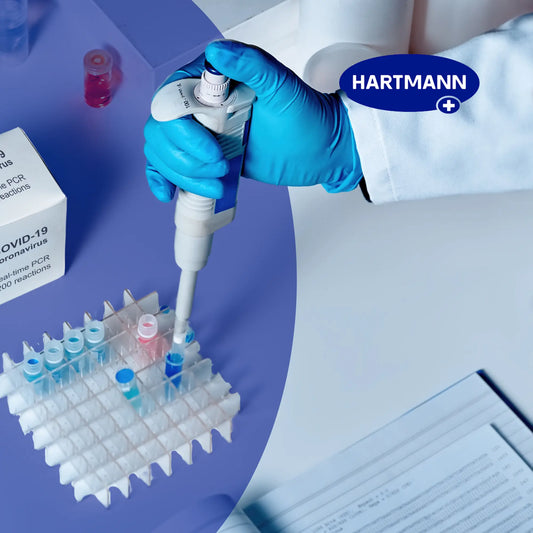 Eine behandschuhte Hand benutzt eine Pipette, um in einem Labor Flüssigkeit in Reagenzgläser zu füllen. Zu sehen sind ein COVID-19-Testkit, ein Reagenzglasgestell, ein Arbeitsblatt und das Wort "HARTMANN".