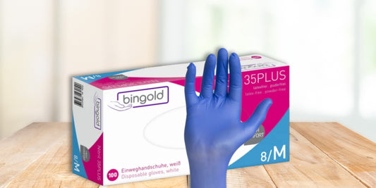 Eine Hand mit blauen Handschuhen wird vor einer Schachtel mit der Aufschrift "bingold 35PLUS" Einweghandschuhe erhoben, die auf einer hölzernen Fläche mit einem unscharfen Hintergrund in einem Innenraum steht.
