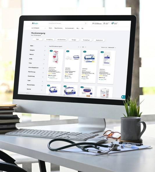 Auf einem Computermonitor wird eine Website für medizinischen Bedarf angezeigt, auf der Produkte zur Wundversorgung vorgestellt werden. Auf dem Schreibtisch neben dem Monitor liegen ein Stapel Notizbücher, ein Stethoskop und eine Topfpflanze.