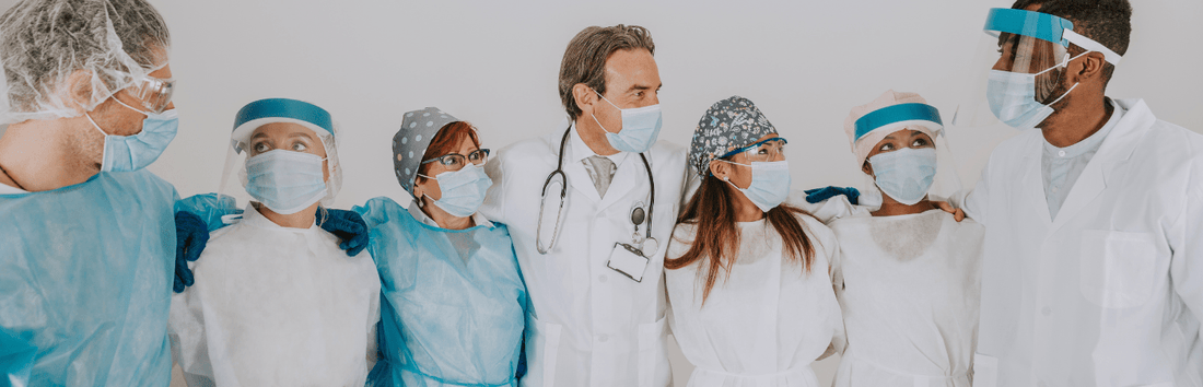 Gruppe von medizinischem Personal mit Schutzkleidung und Masken steht nebeneinander und schaut sich an, in heller Umgebung