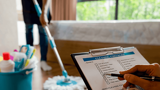 Person hakt Checkliste auf Klemmbrett ab, im Hintergrund wird ein Raum mit Mopp und Reinigungsmitteln gründlich gereinigt