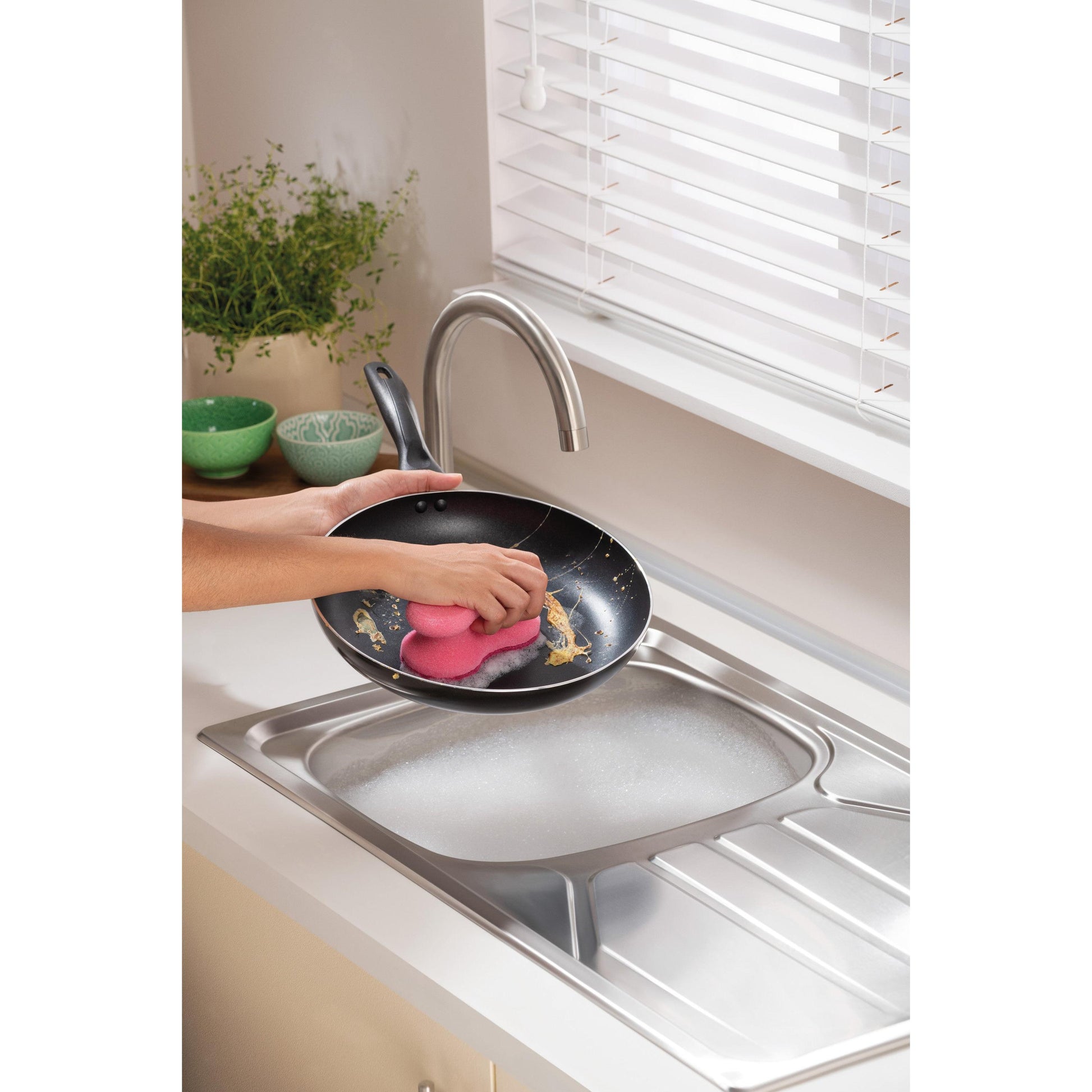Eine Person reinigt eine schmutzige Bratpfanne mit einem rosa Scotch-Brite® Delicate Color Comfort nicht kratzender Reinigungsschwamm von 3M Deutschland GmbH unter einem Wasserhahn in einer seifigen Küchenspüle. Grüne Schalen und eine Topfpflanze stehen auf der Arbeitsplatte am Fenster.