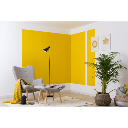 Ein modernes Wohnzimmer mit einem grauen Sessel, einer Ottomane, einer schwarzen Stehlampe, einer gelben Decke und einer Topfpflanze. Die kräftigen gelben geometrischen Formen an der Wand wurden mit 3M Deutschland GmbH ScotchBlue™ Malerabdeckband für scharfe Linien erstellt.