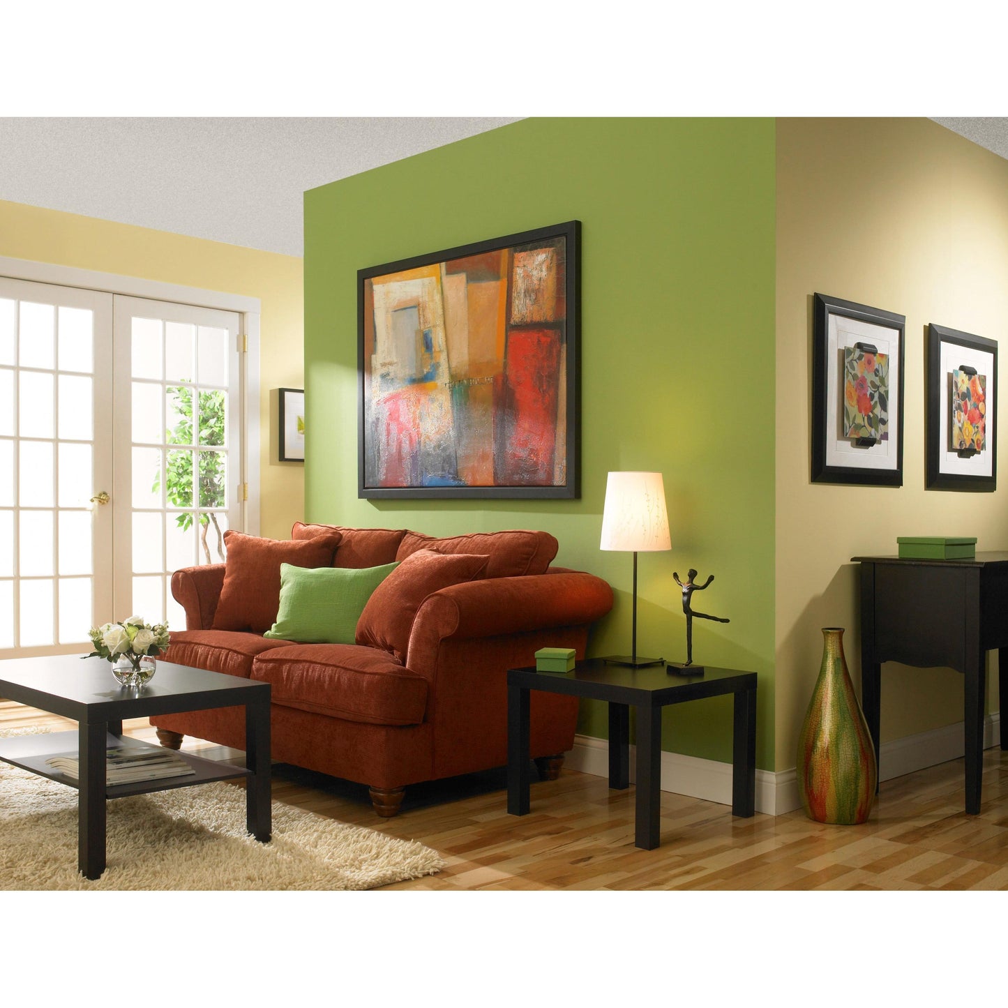 Un soggiorno con pareti gialle e verdi dipinte con nastro adesivo per pittori ScotchBlue™ "Multi-Surface" di 3M Deutschland GmbH, un divano rosso con cuscini verdi, un tavolino da caffè nero, un tavolino con lampada, arte astratta, pavimento in legno e porte in vetro.