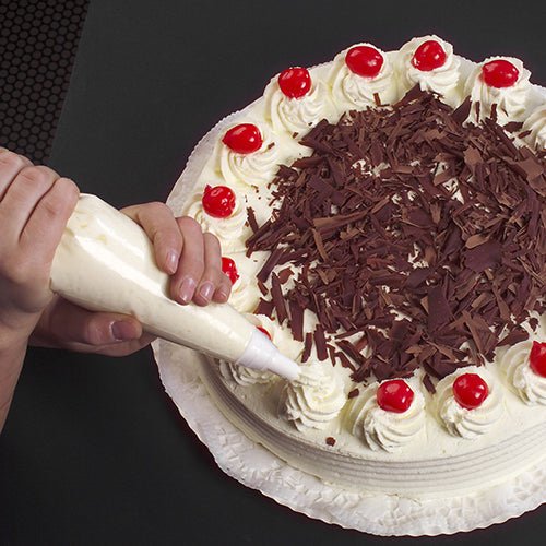 Con il sac à poche Starpak 10 da 1,8 l con 3 bocchette, una torta rotonda viene decorata con panna montata bianca, scaglie di cioccolato al centro e ciliegie sopra, posizionata su un centrino bianco su sfondo scuro.
