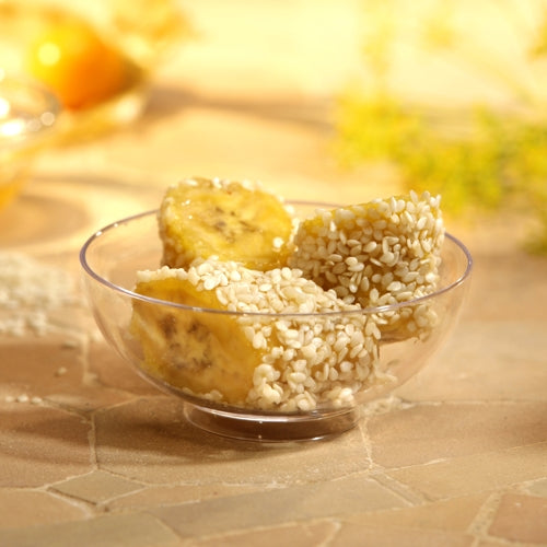 Tre pezzi di banana con semi di sesamo bianco vengono serviti in una ciotola per fingerfood Starpak 50 (PS trasparente), posizionati su una superficie di piastrelle chiare con uno sfondo delicatamente sfocato - ideale per eventi.