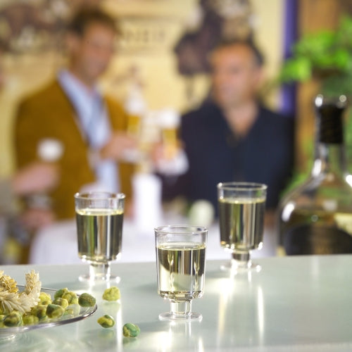 I bicchieri a stelo Starpak 20 per vino bianco in polistirolo trasparente sono su un tavolo con olive e bottiglia, mentre tre persone si intrattengono con bevande sullo sfondo.