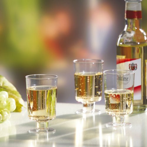 Tre bicchieri a calice Starpak per vino bianco (PS, 0,1l, trasparente) sono riempiti e posizionati accanto a una bottiglia di vino bianco e uva verde su una superficie bianca davanti a uno sfondo sfocato.
