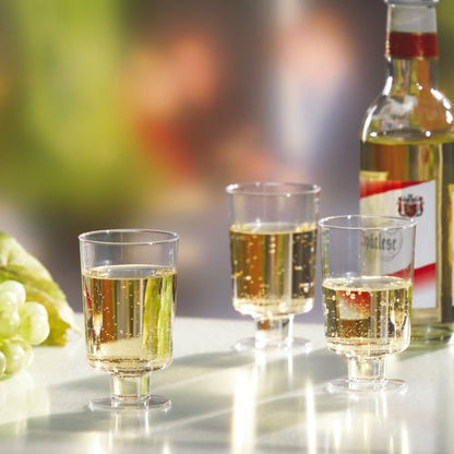 Tre bicchieri a calice Starpak per vino bianco (PS, 0,1l, trasparente) sono riempiti e posizionati accanto a una bottiglia di vino bianco e uva verde su una superficie bianca davanti a uno sfondo sfocato.