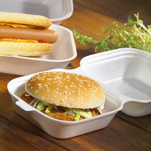 Un hamburger in un panino al sesamo con insalata e polpetta, servito in una scatola per hamburger Starpak 100 con coperchio a cerniera, XPS, 7,5 x 12 x 12 cm bianco - ideale per il catering, con hot dog nelle vicinanze e verdure a foglia su una superficie di legno.