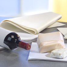 Una bottiglia di vino si trova su una superficie grigia, accanto PAPSTAR sostituto della pergamena "puro" carta da regalo bianca (12,5 kg, resistente ai grassi della PAPSTAR GmbH) e due blocchi di formaggio, uno dei quali parzialmente avvolto. Lo sfondo è leggermente sfocato.