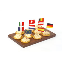 Un vassoio di legno contiene sette cracker, ciascuno con una crema spalmabile e coronato con PAPSTAR decorazioni a stuzzicadenti 8 cm "Nazioni" (vari paesi e UE). Perfetto come decorazione da tavola per feste ed eventi. Marca: PAPSTAR GmbH.