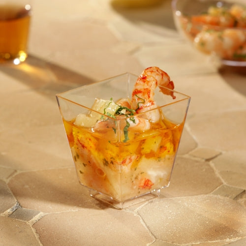 Una ciotola per fingerfood Starpak 50 (PS trasparente) di Starpak, riempita con cocktail di gamberetti e frutti di mare e guarnita con erbe, è posizionata su una superficie strutturata beige. Altre tazze sono sfocate sullo sfondo - perfette per eventi eleganti.