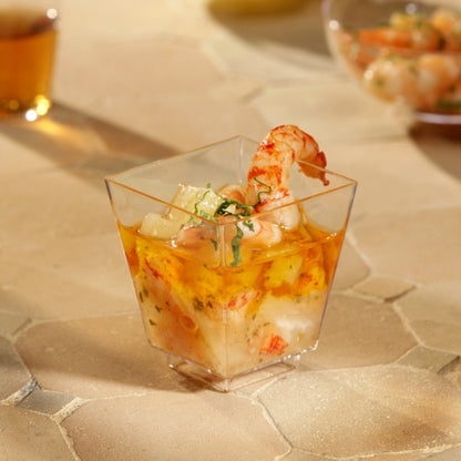 Una ciotola per fingerfood Starpak 50 (PS trasparente) di Starpak, riempita con cocktail di gamberetti e frutti di mare e guarnita con erbe, è posizionata su una superficie strutturata beige. Altre tazze sono sfocate sullo sfondo - perfette per eventi eleganti.