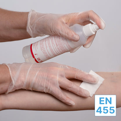 Una persona con guanti monouso sensibili al tatto ARNOMED VINYL PF di ARNOWA GmbH utilizza un flacone spray per bagnare un dischetto di cotone e pulire l'avambraccio di un'altra persona. Nell'angolo in basso a destra appare l'etichetta "EN 455".