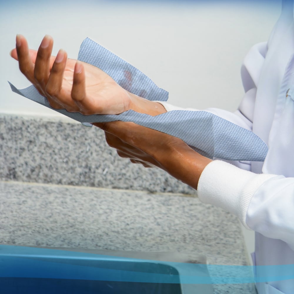 Eine Person in einem weißen Kittel trocknet sich die Hände mit Scott® Handtüchern - Rolle (304,00 m x 19,80 cm) von Kimberly-Clark GmbH von einer Papierhandtuchrolle neben einer Granitarbeitsplatte und einem Waschbecken.