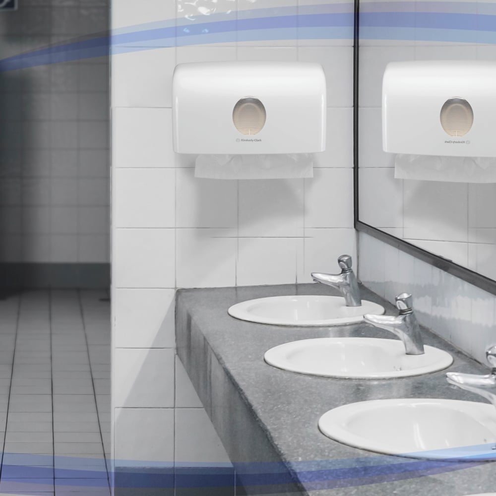 Tre lavandini bianchi con rubinetti argentati si trovano su un piano di lavoro grigio. Sopra, uno specchio e il distributore Aquarius™ per asciugamani piegati di Kimberly-Clark GmbH (MultiFold/Bianco/Piccolo) garantiscono un'estrazione igienica degli asciugamani sulla parete piastrellata.