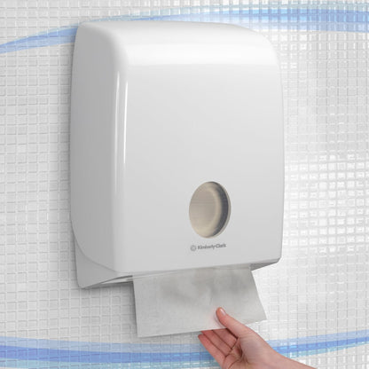 Una mano estrae un asciugamano di carta da un distributore Aquarius™ bianco da parete per asciugamani piegati con piega a C di Kimberly-Clark GmbH, installato su una parete piastrellata.