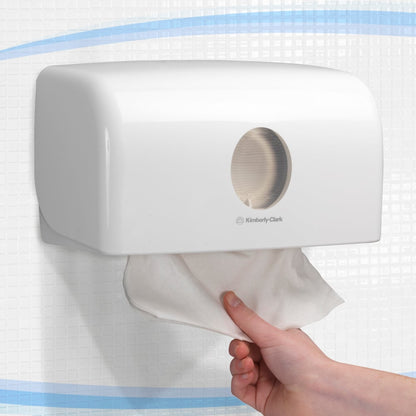 Una mano estrae un asciugamano dal distributore bianco Aquarius™ per asciugamani piegati (MultiFold, piccolo) di Kimberly-Clark GmbH, montato a parete su una parete piastrellata per un'estrazione igienica.