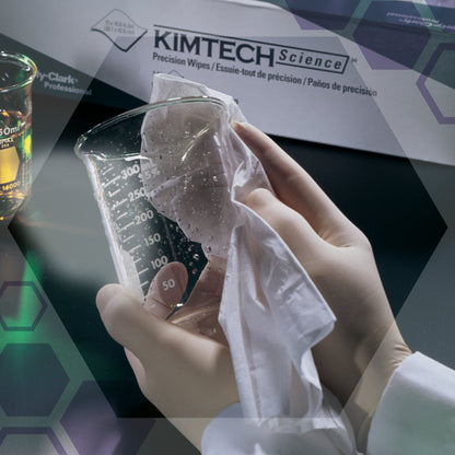 Una persona con guanti bianchi pulisce un bicchiere di vetro trasparente con salviette di precisione Kimtech® Science della Kimberly-Clark GmbH. Sullo sfondo ci sono una scatola con le salviette (100 fogli, 2 strati) e un bicchiere con del liquido.