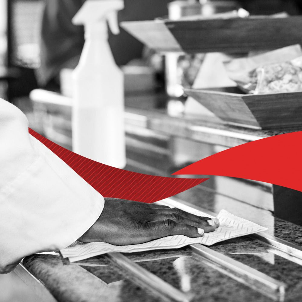 Una mano pulisce un piano di lavoro in granito con un rotolo di carta assorbente WypAll® L20 (2 strati, 6 x 550 fogli) della Kimberly-Clark GmbH; sullo sfondo si vedono una bottiglia spray e ciotole per alimenti, con un elemento grafico rosso che attraversa l'immagine.