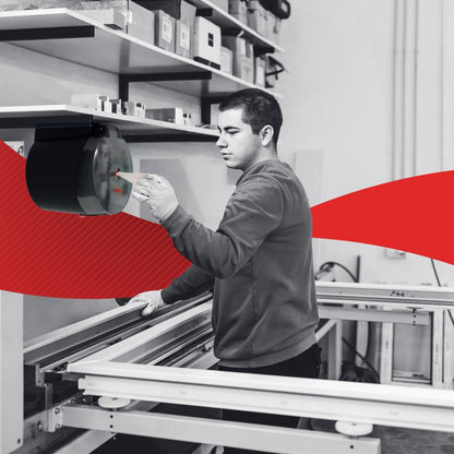 Una persona utilizza un dispenser WypAll® Reach Plus™ Reach™ Centrefeed montato a parete della Kimberly-Clark GmbH in un luogo di lavoro. Dietro ci sono scaffali con cartoni e altri oggetti, mentre una grafica rossa attraversa orizzontalmente l'immagine.