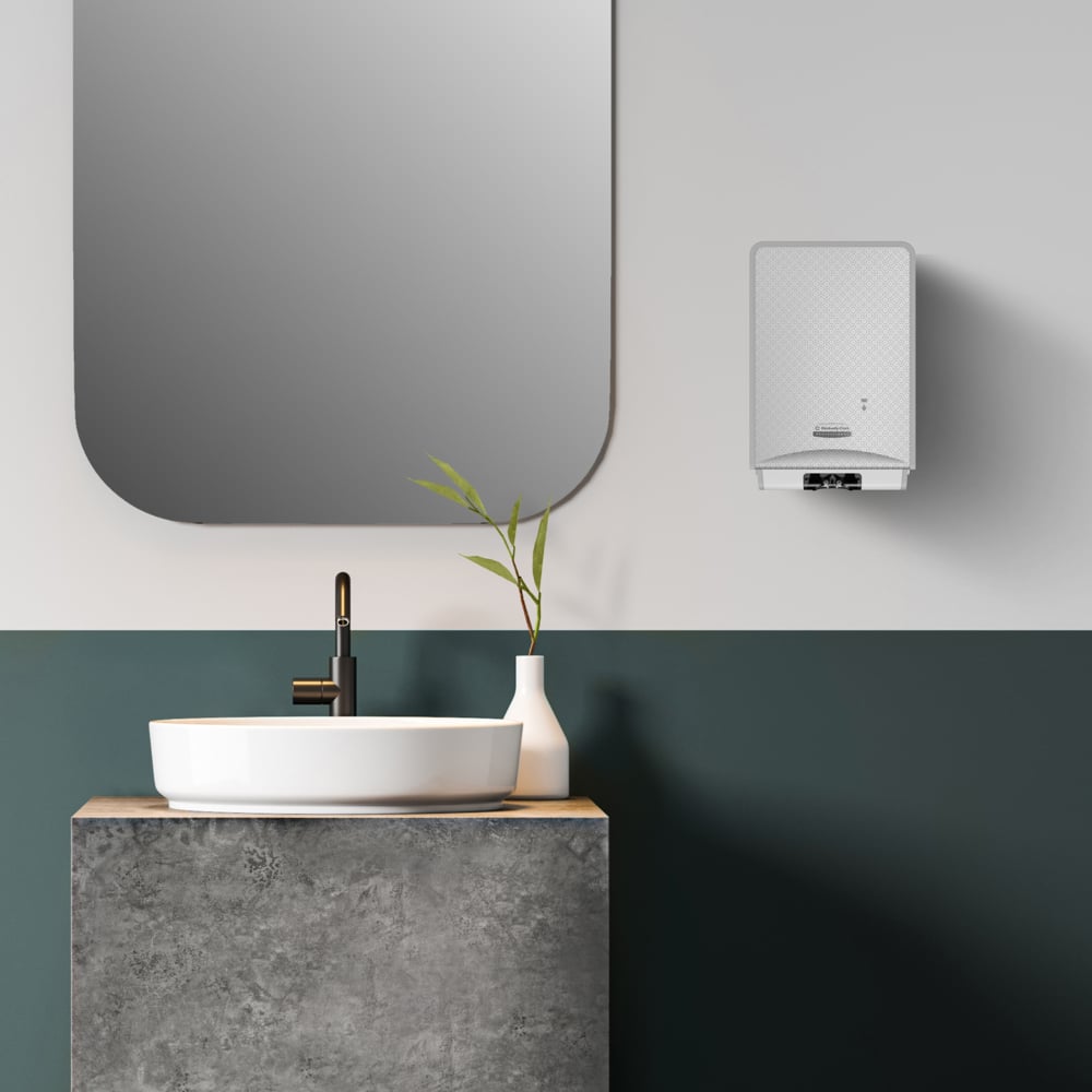 Un bagno moderno con un lavabo rotondo bianco su un piano in cemento, un rubinetto nero, un vaso bianco con steli verdi, un grande specchio rettangolare e una cassetta da 1,2 litri di Kimberly-Clark Kleenex® Botanics™ Luxury Foam Hand Wash montata a parete.