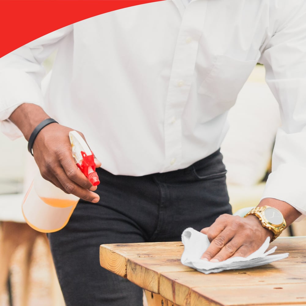 Una persona con camicia bianca e pantaloni neri usa un flacone spray e un rotolo grande WypAll® L40 PowerClean (Kimberly-Clark GmbH) per pulire un tavolo di legno. La persona indossa un orologio; il bordo superiore dell'immagine mostra una cornice rossa ondulata.