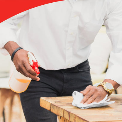 Una persona con camicia bianca e pantaloni neri usa un flacone spray e un rotolo grande WypAll® L40 PowerClean (Kimberly-Clark GmbH) per pulire un tavolo di legno. La persona indossa un orologio; il bordo superiore dell'immagine mostra una cornice rossa ondulata.