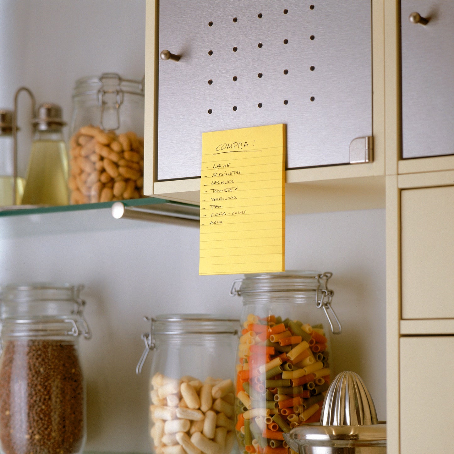 Ein handgeschriebener Einkaufszettel in spanischer Sprache befindet sich auf einem beigen Küchenschrank unter Verwendung eines 3M Deutschland GmbH Post-it® Super Sticky Notes im Großformat, Ultra Gelb (101 x 152 mm), über Glasgefäßen mit Nudeln, Bohnen und Getreide. In den benachbarten Regalen befinden sich weitere Gläser und Küchenutensilien.