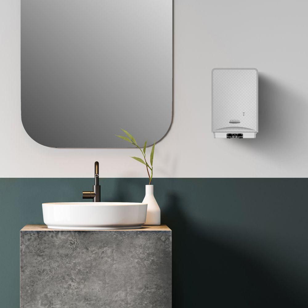 Un bagno moderno dispone di uno specchio rotondo, un lavandino in ceramica bianca, un rubinetto nero e il Detergente Schiuma per le Mani Scott® Control™ (4 cartucce) di Kimberly-Clark GmbH per un'igiene ottimale delle mani. Accanto al lavandino c'è un vaso con foglie verdi, alla sua destra si trova un scaldabagno.