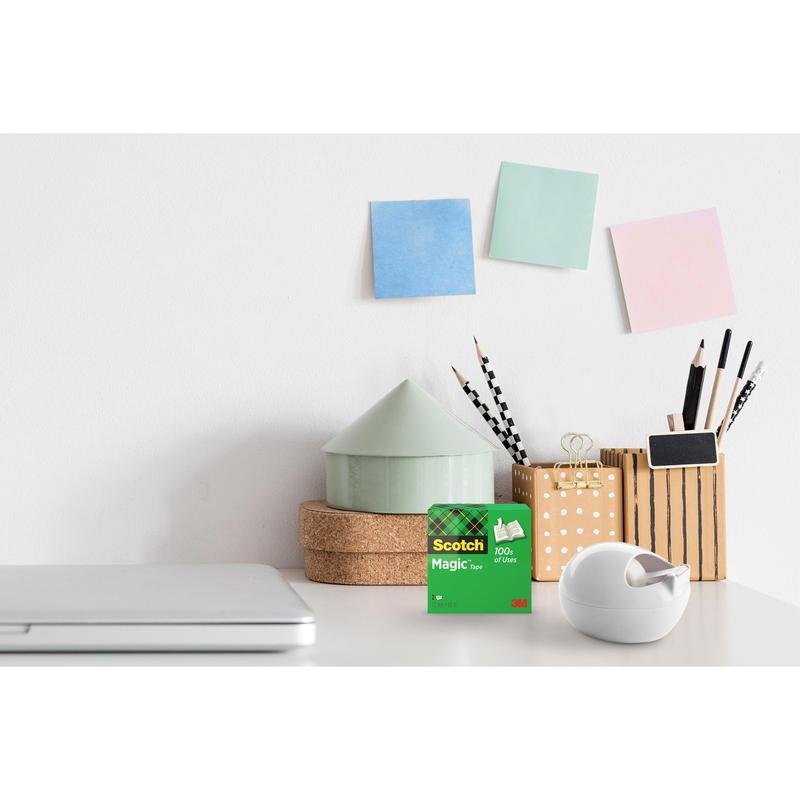 Una scrivania con un laptop chiuso, un dispenser da tavolo Scotch™ Karim Rashid di 3M Deutschland GmbH e una scatola di nastro adesivo, contenitori per penne e matite, un piccolo ornamento a forma di casa e tre note adesive colorate sulla parete bianca.
