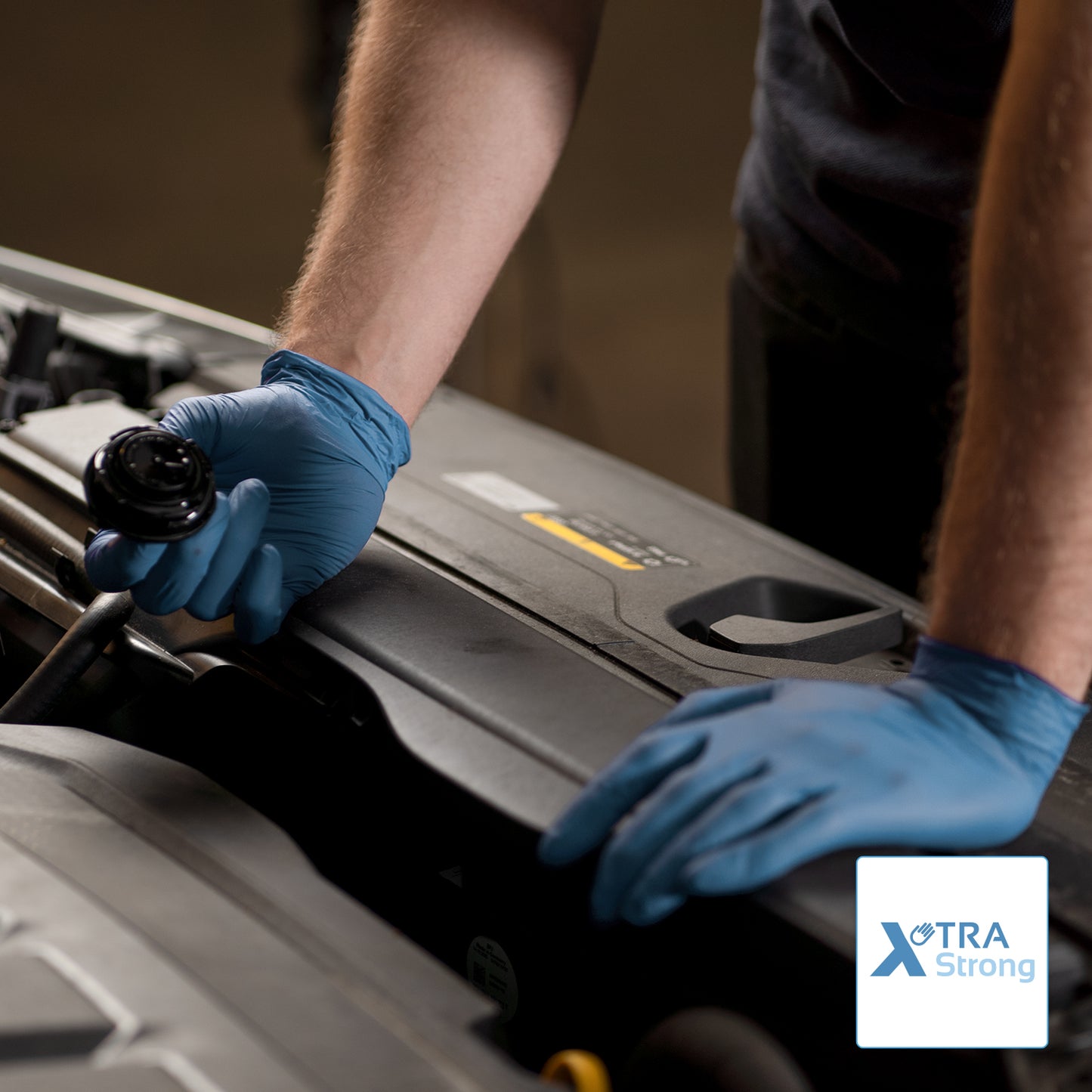 Una persona con guanti monouso ARNOMED NITRIL BLUE XTRA STRONG di ARNOWA GmbH apre il cofano di un'auto. Si vedono solo il busto e le braccia, in basso a destra è raffigurato il logo Xtra Strong.