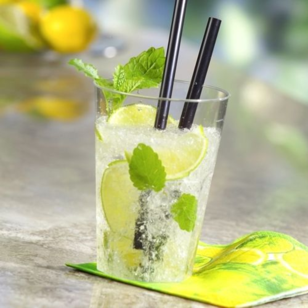 Un bicchiere di plastica riutilizzabile Starpak, riempito con un cocktail di lime e menta, guarnito con fette di lime fresco e foglie di menta. All'interno ci sono due cannucce nere, posizionate su un tovagliolo con motivo a limone. Lo sfondo sfocato mostra altri lime e piante verdi.