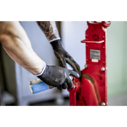 Una persona con guanti in nylon NITRAS, neri, rivestiti in PU di AS Arbeitsschutz GmbH pulisce o lubrifica con una bomboletta spray blu una parte di macchina rossa in metallo. Si vedono solo braccia e mani.