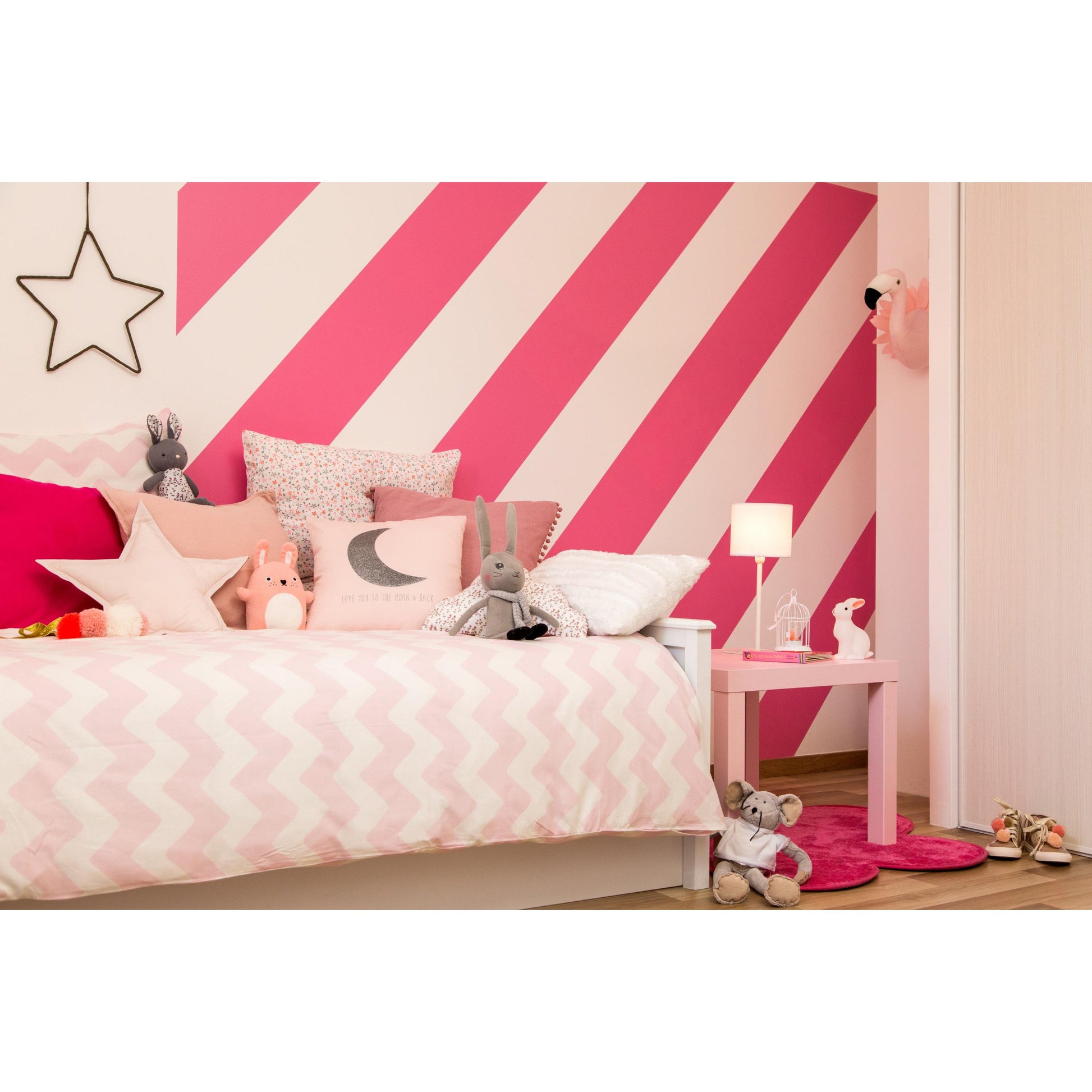 Ein Kinderzimmer mit weißem Bett, rosa und grauen Kissen, Plüschtieren und kräftigen rosa-weiß diagonal gestreiften Wänden, gestrichen mit ScotchBlue™ Malerabdeckband für scharfe Linien von 3M Deutschland GmbH. Eine Lampe, ein Tisch, Spielzeug und Schuhe vervollständigen den gemütlichen Raum.