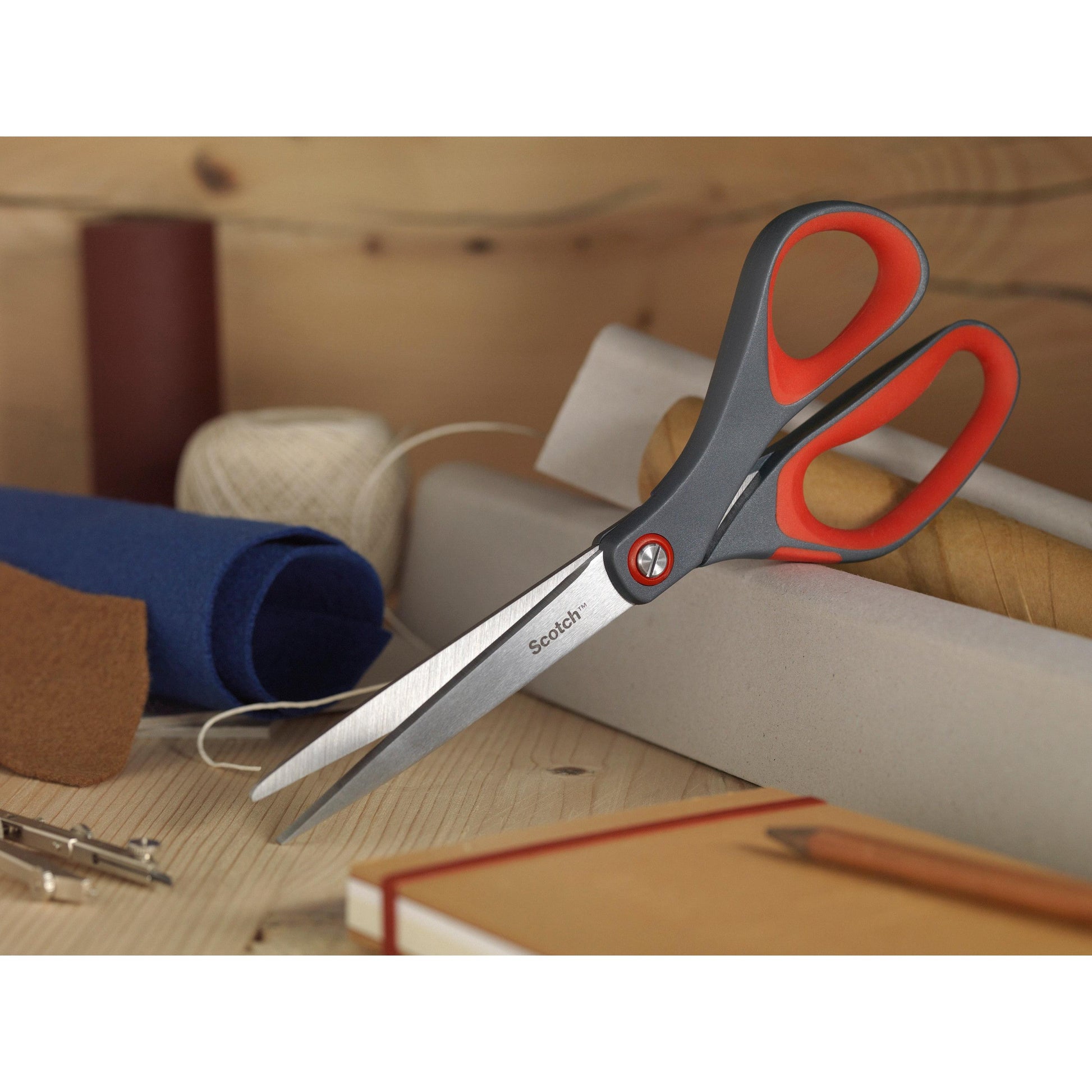 Die Scotch™ Präzisionsschere rot von 3M Deutschland GmbH mit grauem und rotem Griff liegt auf einer Holzfläche zwischen Stoffrollen, einer Spule mit weißem Garn, einem braunen Notizbuch und einem Bleistift.