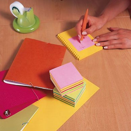 Su una scrivania di legno, qualcuno scrive con una penna arancione su un blocco di Post-it® Notes rosa (76 mm x 76 mm, 450 fogli/blocco, 100% PEFC) della 3M Deutschland GmbH. Vicino ci sono un taccuino rosso, un dispenser di nastro adesivo verde, una campanella argentata e altri foglietti colorati.