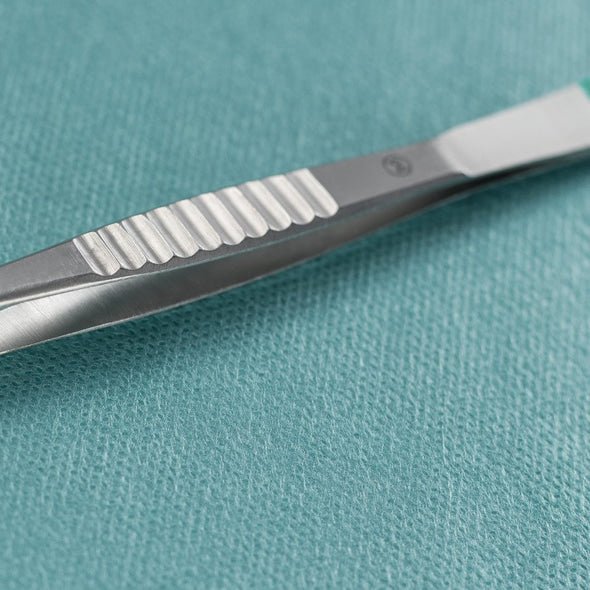 Un primo piano di una pinzetta Hartmann Splitter della Paul Hartmann AG dritta 9cm, sterile (confezione da 40 pezzi), che si trova accanto a un bisturi chirurgico in metallo su un panno medico strutturato color teal.