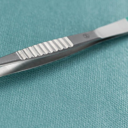 Un primo piano di una pinzetta Hartmann Splitter della Paul Hartmann AG dritta 9cm, sterile (confezione da 40 pezzi), che si trova accanto a un bisturi chirurgico in metallo su un panno medico strutturato color teal.