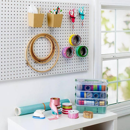 Un'area di lavoro artigianale con una bacheca a cui sono appese le forbici di precisione rosse Scotch™ di 3M Deutschland GmbH, cerchi da ricamo, rotoli di nastro adesivo e penne. Sotto ci sono materiali artigianali e nastri su un tavolo bianco vicino a una finestra.