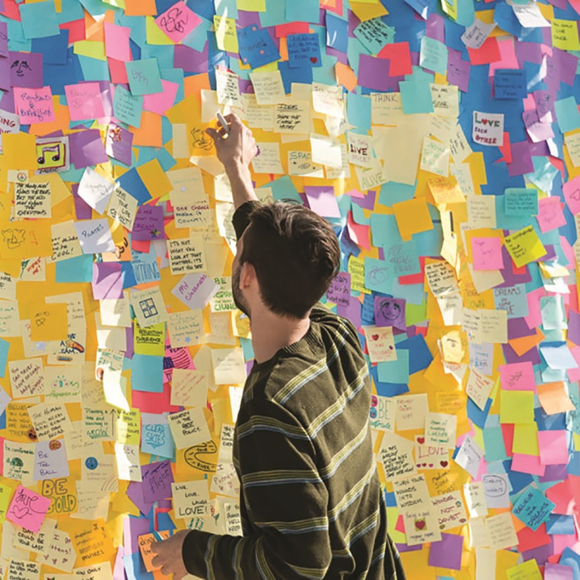 Eine Person im gestreiften Hemd klebt einen Zettel an eine Wand, die mit bunten 3M Deutschland GmbH Post-it® Notes Würfel, Ultra-Farben (76x76 mm, 450 Blatt/Block) gefüllt ist, die jeweils handschriftliche Nachrichten und Zeichnungen zeigen.