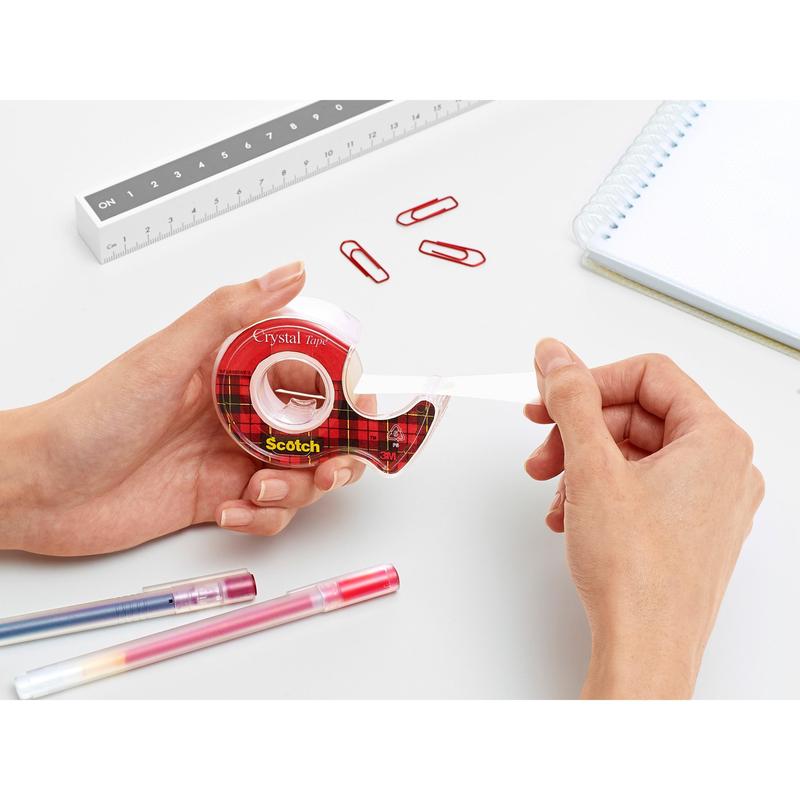Una persona tira il nastro adesivo Scotch® Crystal (19 mm x 7,5 m) dal dispenser rosso della 3M Deutschland GmbH su una scrivania bianca con graffette rosse, penne, un righello e un quaderno a spirale.