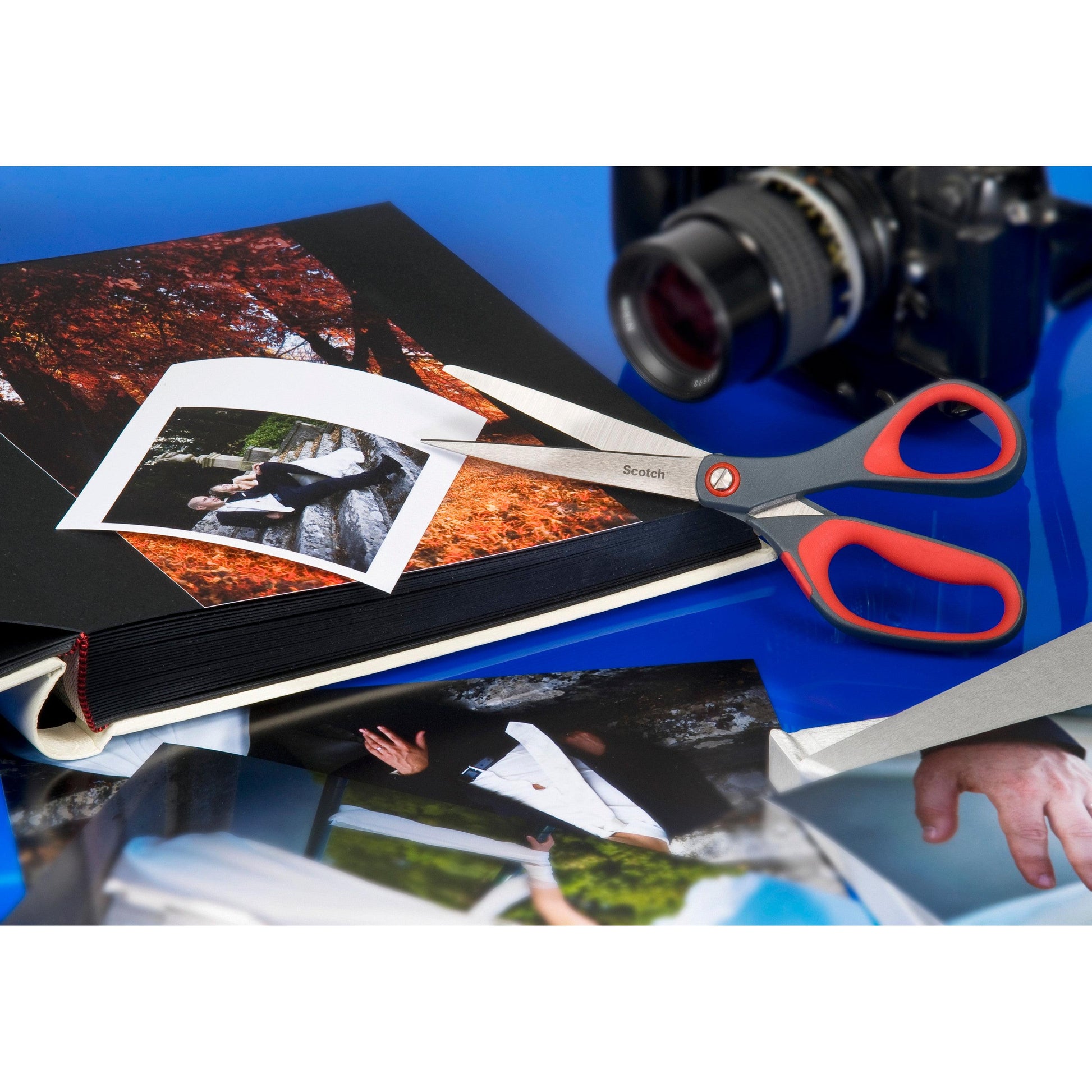 Eine Scotch™ Präzisionsschere rot von 3M Deutschland GmbH liegt auf einem geöffneten Fotoalbum, eine lose Fotografie ist teilweise geschnitten. Daneben befinden sich weitere Fotos und eine Kamera auf einer blauen Fläche.