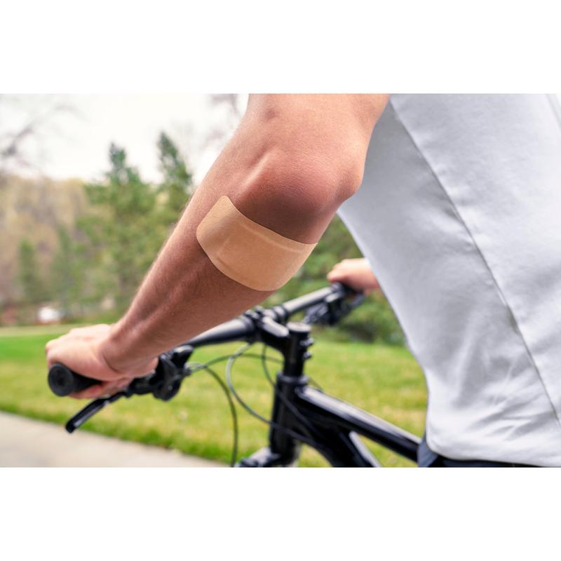 Una persona che indossa una camicia bianca e va in bicicletta all'aperto ha un cerotto Nexcare™ Ultra Stretch MAXI Comfort Flexible (50 mm x 101 mm) di 3M Deutschland GmbH sul braccio nudo; sullo sfondo si vedono erba verde e alberi.