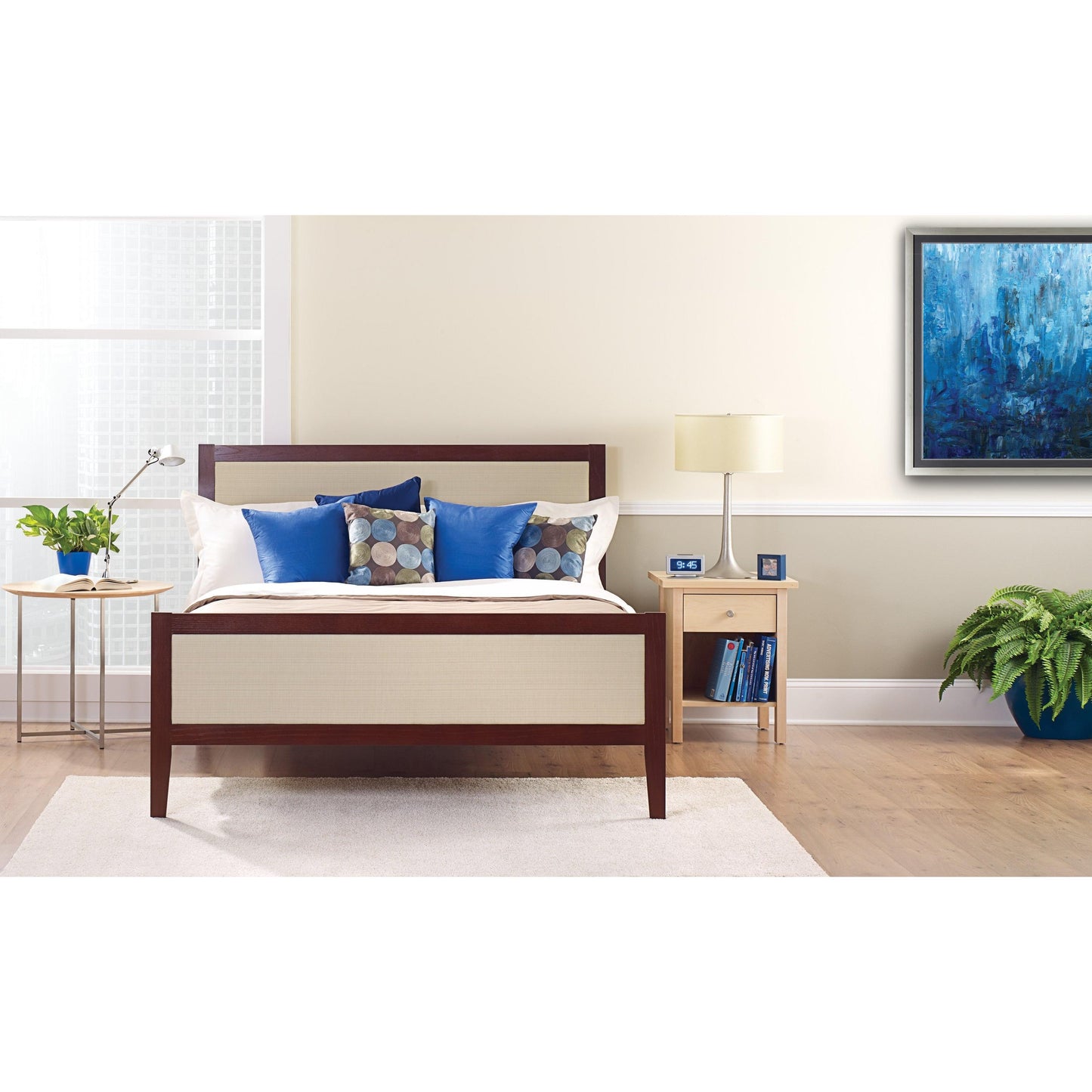 Un letto ben fatto con cuscini bianchi e blu si trova davanti a una parete chiara, coperta con il nastro adesivo per pittori ScotchBlue™ "Multi-Surface" della 3M Deutschland GmbH. Su un comodino ci sono una lampada, un orologio, libri e una pianta in vaso accanto alla finestra luminosa.