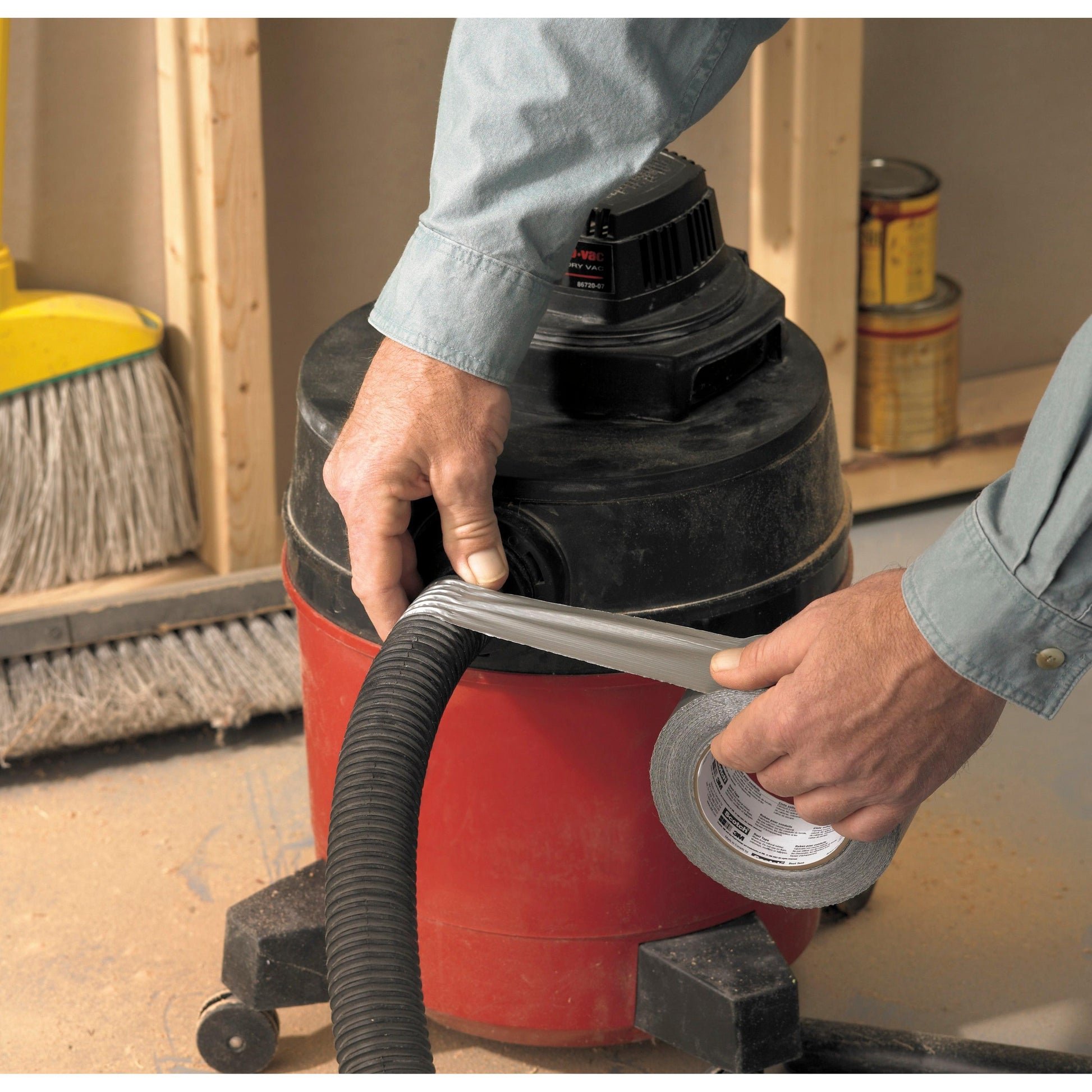 Eine Person wickelt Scotch® Universal Klebeband 2904 (50 m x 48 mm) von 3M Deutschland GmbH um einen Staubsaugerschlauch, der an einem roten Werkstattsauger befestigt ist. Im Hintergrund sind ein Besen und Farbdosen zu sehen.