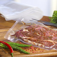 Una porzione di carne marinata è posta in un PAPSTAR sacchetto sigillato 100 trasparente (PA/PE) della PAPSTAR GmbH su un tagliere di legno con peperoncini e grani di pepe, sottolineando così la conservazione degli alimenti. Sullo sfondo si vedono verdure a foglia.